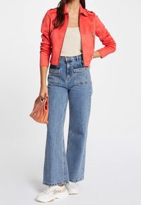 Czerwona kurtka z sztucznego zamszu, beżowa top, wysokie niebieskie dżinsowe spodnie typu wide-leg, białe trampki i wzorzona pomarańczowa torebka.