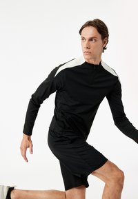 Sort atletisk langærmet top med hvide skulderdetaljer og høj lynlås krave, parret med sorte shorts. Glat, strækbart stof.