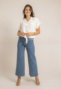 Chemisier blanc à manches courtes avec col et détails de boutons, assorti à un jean en denim bleu à taille haute et jambes larges, avec des boutons dorés accentués.