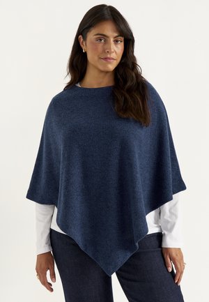 Woman with long dark hair wearing a navy blue poncho over a white shirt and dark pants standing against a plain light background.