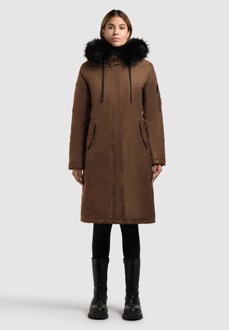 Brun polstret frakke med hætte i sort kunstpels, snor i taljen og to forlommer. Båret over sorte leggings og kraftige sorte støvler.