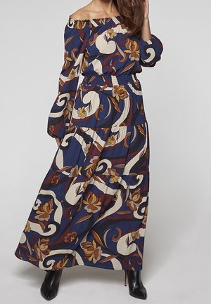 Femme portant une robe longue bleu marine épaules dénudées avec des motifs floraux et abstraits marron, crème et jaune, associée à des bottes noires à talons.