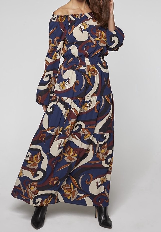 Femme portant une robe longue bleu marine épaules dénudées avec des motifs floraux et abstraits marron, crème et jaune, associée à des bottes noires à talons.