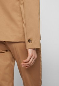 Ręka wyluzowana obok beżowych spodni garniturowych i rękawa marynarki z pojedynczym brązowym guzikiem przy mankiecie, na jednolitym tle.