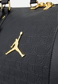 Svart teksturert duffelbag med et gyllent Jumpman-logo og preget '23'-mønster, med gyllent hardware og doble svarte stropper.