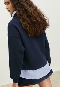 Mörkblå oversized sweatshirt med strukturerad yta, lager på lager över en blå och vit randig skjorta. Har rundad nederkant och sänkta axlar.