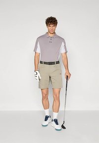 Hombre en polo de golf gris, pantalones cortos beige, guante de golf blanco y zapatos blancos sosteniendo un palo de golf contra un fondo blanco.