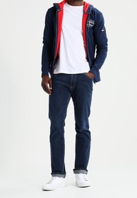 Felpa con cappuccio blu navy con interno rosso, logo bianco, abbinata a una t-shirt bianca e jeans blu scuro. Scarpe da ginnastica bianche completano il look.