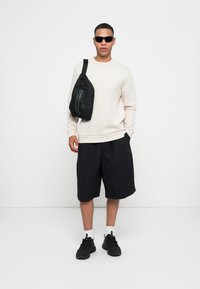 Sudadera beige clara, pantalones cortos negros, bolso cruzado negro y zapatillas negras. Los accesorios incluyen gafas de sol oscuras y calcetines blancos.