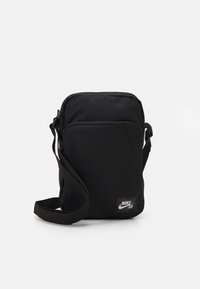 Borsa a tracolla nera in nylon con cinturino regolabile, chiusura a zip e una piccola toppa del logo Nike SB sul davanti. Forma rettangolare.
