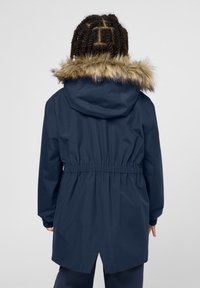 Cappotto impermeabile blu navy con vita regolabile tramite coulisse, cappuccio foderato in pelliccia e polsini elasticizzati. La vista posteriore mostra un taglio più lungo e un dettaglio dell'orlo spaccato.