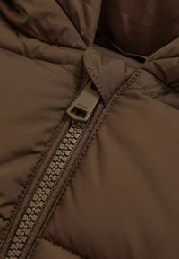 Brūna pūkainā jaka ar rāvējslēdzēju, uz kura ir zīmola nosaukums "REISS" uz vilkšanas cilpas. Apaļš apkakle un quilted tekstūra.