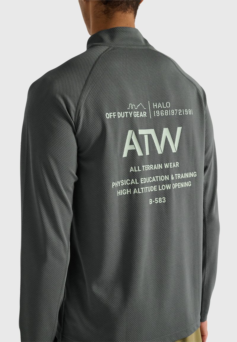 Mann trägt graues, strukturiertes Langarmshirt mit der Aufschrift "ATW All Terrain Wear" und zusätzlichem Text auf der Rückseite.