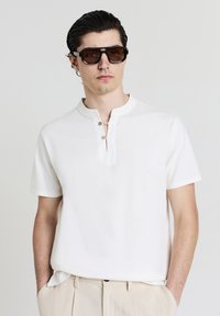 Uomo con capelli scuri che indossa grandi occhiali da sole rettangolari, una camicia bianca con maniche corte e pantaloni beige, con le mani in tasca.