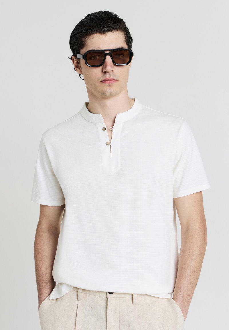 Uomo con capelli scuri che indossa grandi occhiali da sole rettangolari, una camicia bianca con maniche corte e pantaloni beige, con le mani in tasca.