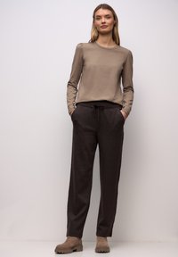 Frau trägt taupefarbenes Langarmshirt, dunkelbraune Hose mit Kordelzug und taupefarbene Stiefeletten, steht vor einem schlichten hellen Hintergrund.