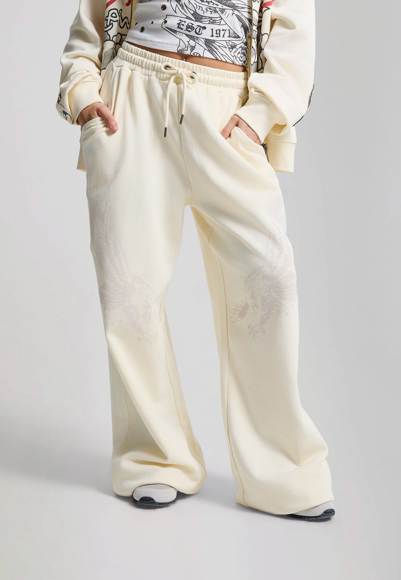 Person wearing loose cream sweatpants with faint dragon prints, white graphic cropped top, and matching cream jacket with hands in pockets.