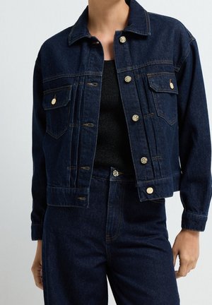 Veste en denim bleu foncé avec des boutons dorés portée sur un haut noir, associée à un pantalon en denim bleu foncé assorti.