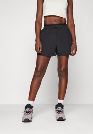 Athletic person wearing black drawstring shorts, white crop top, white socks, and light purple athletic shoes standing on plain background.