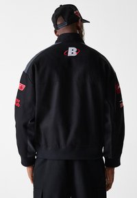 Black jacket with grey accents, ribbed cuffs and hem, featuring logos in red on sleeves and back. Durable fabric with a relaxed fit.