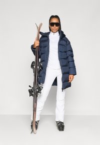 Jachetă puffer navy, bluză albă cu fermoar, pantaloni de schi albi, ținând schiuri. Design elegant, textură pufoasă în jachetă, echipată cu brățări pentru schi.