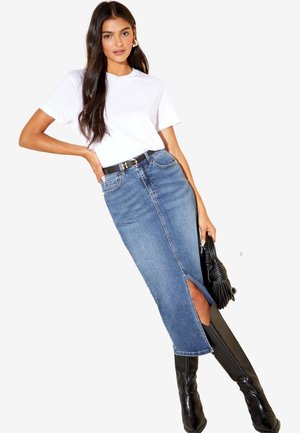 Femme aux longs cheveux noirs portant un t-shirt blanc, une jupe midi en denim bleu avec une fente à l'avant, une ceinture noire, des bottes noires montantes et tenant un sac à main noir.