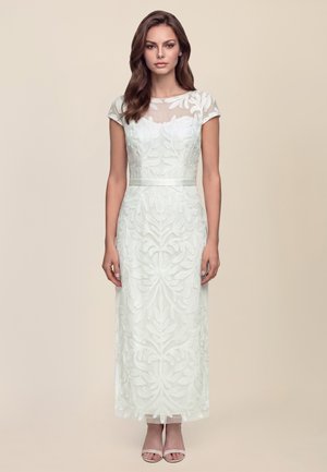 Woman stands wearing a white, embroidered ankle-length dress with short sleeves and sheer neckline, paired with white open-toe heels.