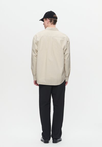 Chemise beige à manches longues avec un col boutonné, portée avec un pantalon noir. La chemise présente un tissu texturé et une coupe droite.