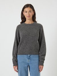 Mulher com cabelo castanho, usando brincos de argola dourados, um suéter cinza escuro e jeans azuis claros, de pé contra um fundo branco liso.