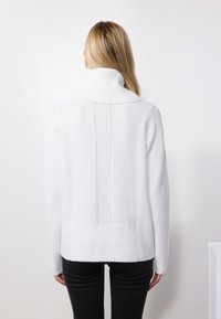 Maglione bianco a collo alto con colletto e orlo a costine, caratterizzato da un design testurizzato sul retro. Indossato con pantaloni scuri.
