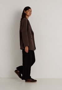 Barna, túlméretezett blézer két gombbal, fekete, bővített szárú nadrág és barna, textúrázott részletekkel ellátott sneakers. A modell profilban áll, fehér háttér előtt.