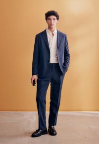 Giovane uomo in abito blu a righe sottili, camicia bianca e mocassini neri, in piedi contro una parete beige, con occhiali da sole nella mano destra.