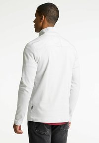 Hvid langærmet pullover med ståkrave, der har en glat tekstur og sideslids, set bagfra. Tynde syningsdetaljer er synlige.