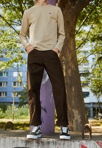 Beige Langarmhemd mit rotem Logo, dunkelbraune Hose und schwarz/weiße gemusterte Sneakers. Das Modell steht gegen einen Baum mit urbanem Hintergrund.