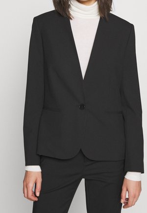 Vrouw draagt een nauwsluitend zwart colbert, één keer dichtgeknoopt, over een witte coltrui en bijpassende zwarte broek, staand tegen een effen achtergrond.