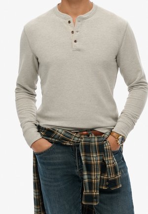 Mann trägt hellgraues langärmeliges Henley-Shirt, blaue Jeans, kariertes Hemd um die Taille gebunden und eine Armbanduhr mit braunem Lederarmband.