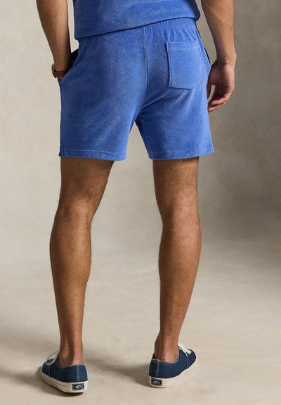 Άνδρας που φοράει μπλε σορτς από πετσετέ ύφασμα και ταιριαστό τοπ, σκούρα μπλε αθλητικά παπούτσια με λευκές σόλες, στέκεται με τα χέρια χαλαρά δίπλα στο σώμα.