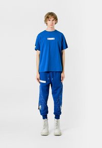 Camiseta de algodón azul con un parche rectangular blanco en el frente; combinada con pantalones jogger azules que presentan intrincados patrones en forma de plumas y acentos blancos.