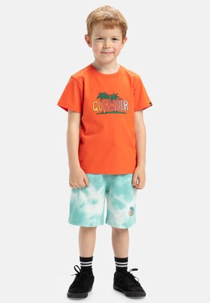 Ljusa orange bomullströja med palmträdsgrafik, ihopparad med teal tie-dye shorts. Svarta skor med vita ränder på strumporna.