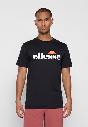 Mann trägt schwarzes ellesse-T-Shirt mit weißem und orangefarbenem Logo, kombiniert mit rötlich-braunen Shorts, stehend vor einem einfarbigen hellgrauen Hintergrund.