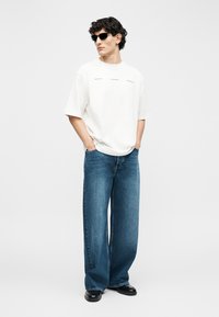 Férfi fekete napszemüvegben, túlméretezett fehér pólóban, bőszárú kék farmerben és fekete cipőben, zsebekbe tett kézzel áll.