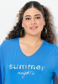 Modrá bavlněná tričko s V-výstřihem, na kterém jsou slova "letní noci" v barevném květinovém tisku. Textura, volný střih s krátkými rukávy.