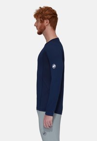 Marineblauwe longsleeve shirt in een getailleerd ontwerp, gemaakt van rekbare stof met een ronde hals en een embleem op de linkermouw. Grijze broek zichtbaar.