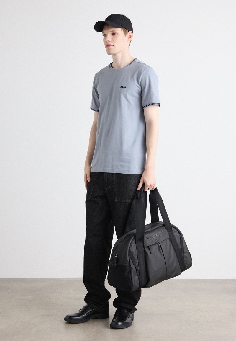 T-shirt gris à manches courtes avec un logo, pantalons noirs, casquette noire et un sac de sport noir avec une poche frontale et des poignées. Tissu lisse.