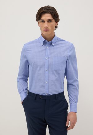 Young man wearing a light blue button-up shirt with chest pocket and dark blue trousers, standing with one hand in pocket against plain background.