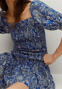 Blaues Kleid mit Paisleymuster, kurzen Puffärmelchen, quadratischem Ausschnitt und geraffter Taille. Hergestellt aus leichtem Stoff mit floralen Akzenten.