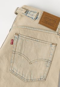 Πίσω τσέπη μπεζ τζιν Levi's που δείχνει κόκκινη ετικέτα Levi's και καφέ δερμάτινο λογότυπο Levi Strauss & Co. πάνω από τη μέση.