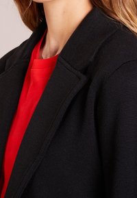 Svart texturerad kavaj med krage, lagd över en röd skjorta. Har en skräddarsydd silhuett och mjukt tyg som framhäver rena linjer.