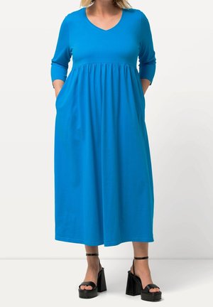 Femme portant une robe midi bleu vif à manches longues avec poches, associée à des sandales plateformes noires avec brides à la cheville.