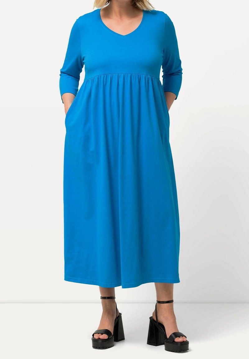 Femme portant une robe midi bleu vif à manches longues avec poches, associée à des sandales plateformes noires avec brides à la cheville.